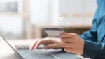 person sitting in front of laptop holding a bank card with a error sign indicating fraud