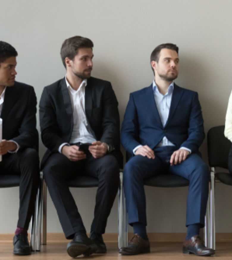 Candidates seated in line for an interview, with a woman in business attire holding a resume and engaging with a male applicant