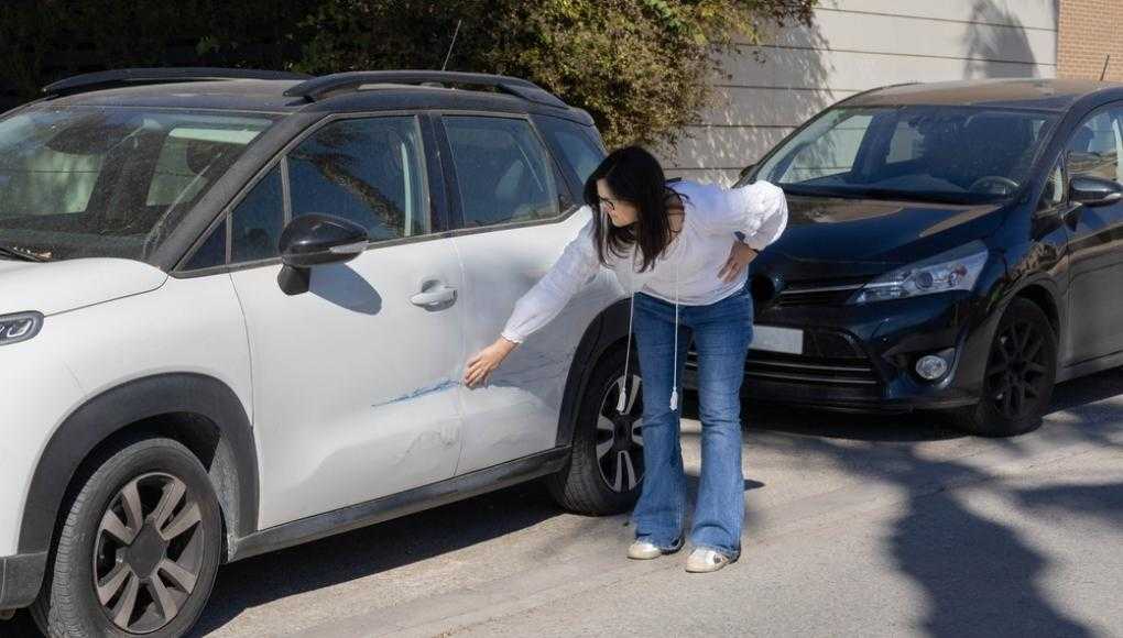 Woman inspecting scratch on white car's door in parking lot, potential car accident claim scenario.