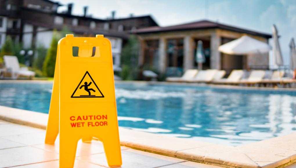 Yellow caution wet floor sign near the edge of a swimming pool at a resort, with lounge chairs and a building in the background