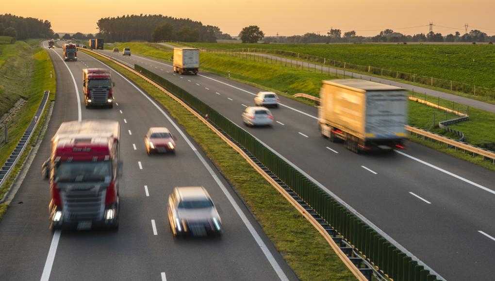 Busy highway traffic at sunset with fast-moving cars and trucks on a divided road, surrounded by lush green fields