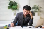 person sitting at desk with piles of papers, laptop nearby
