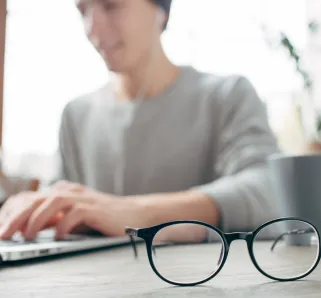 Cut view of a guy sitting at the table and wokring. He is wokring using the laptop. Besides the laptop there is a cup of tea. There are glasses in the front of the picture