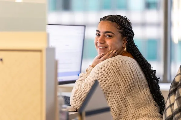 Smiling woman with braided hair and a beige sweater sits at a desk