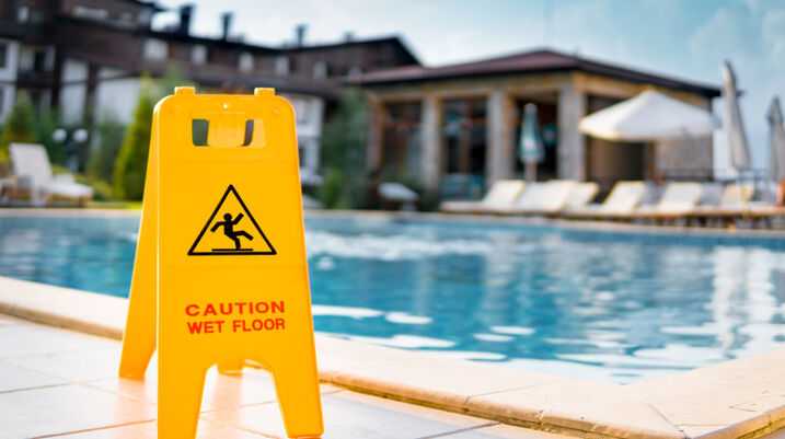 Yellow caution wet floor sign near the edge of a swimming pool at a resort, with lounge chairs and a building in the background