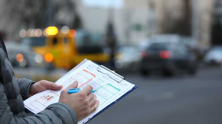 Person filling out an accident report form on a clipboard with blurred traffic and a tow truck in the background