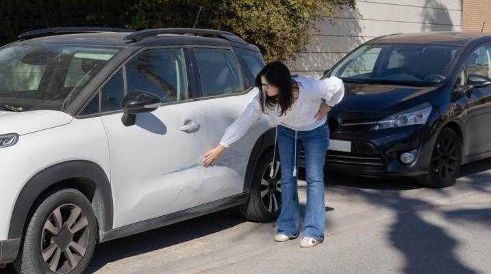 Woman inspecting scratch on white car's door in parking lot, potential car accident claim scenario.