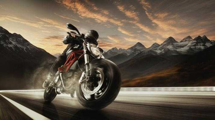 Motorcyclist speeding on a scenic mountain road at sunset with dramatic sky and mountain backdrop.