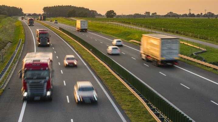 Busy highway traffic at sunset with fast-moving cars and trucks on a divided road, surrounded by lush green fields