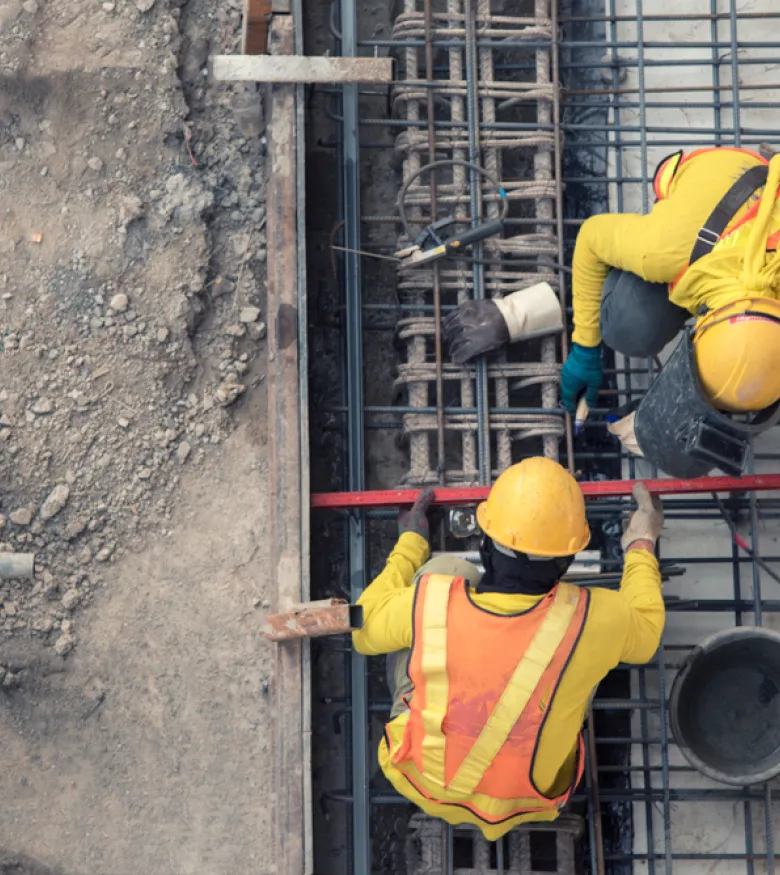 Aerial view of two construction workers in yellow and orange safety vests working on steel reinforcements at a construction site.