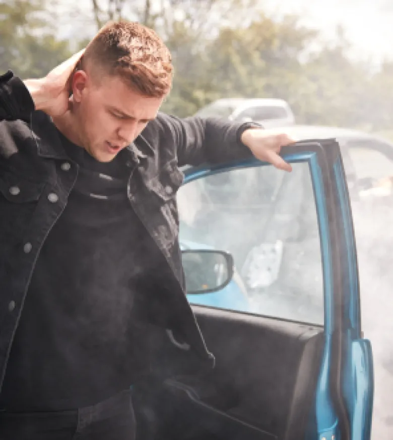 A man stands outside his car holding his neck, looking pained, with airbags deployed inside the vehicle, illustrating Airbag Injuries in The Villages.